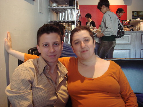 Two people sitting in a coffee shop, both with short dark hair. One is wearing a beige button-down shirt, and the other is wearing an orange scoop-necked shirt.