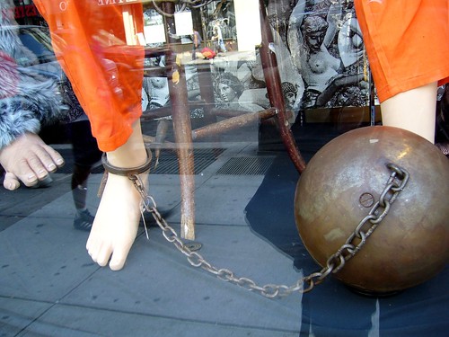 Doll with metal ball cuffed to the legs on shop window.