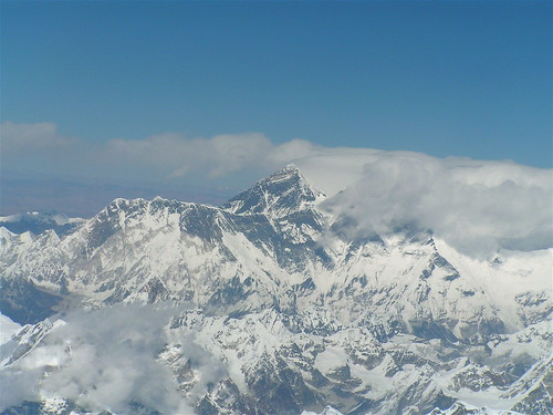A photograph of Mount Everest, viewed from the distance so that other mountains can be seen for size comparison. A dusting of snow is sprinkled across the landscape and mist swirls around the mountain.